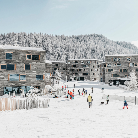 Situé directement à la station de la vallée, le rocksresort à Laax offre le plaisir du ski dès le pas de la porte – design moderne et esprit alpin s’y rencontrent (Photo : © LAAX / Weisse Arena Gruppe) Vue hivernale du rocksresort à Laax avec des skieurs devant une architecture moderne et une forêt enneigée