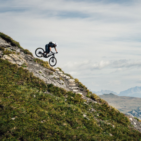Ein Mountainbiker auf einem Bergpfad vor majestätischer Alpenkulisse in Arosa unter blauem Himmel.