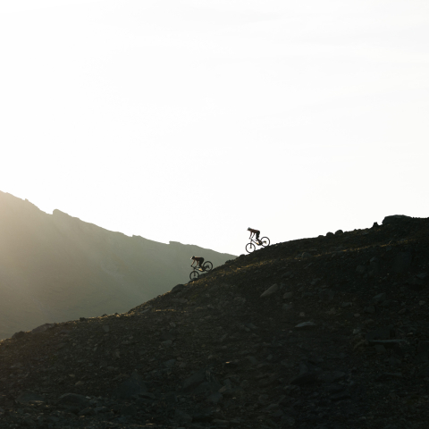 Zwei Mountainbiker auf einem steilen, steinigen Trail in den Bergen. Die Szene ist in das helle Gegenlicht der untergehenden Sonne getaucht.