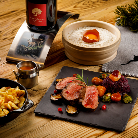 Ein gedeckter Holztisch mit einem Wildgericht auf einer Schieferplatte, serviert mit Spätzli in einer gusseisernen Pfanne, einem Glas Rotwein und herbstlicher Dekoration