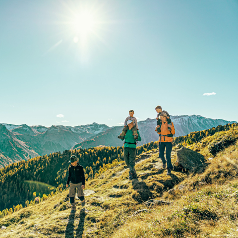 Familie mit drei Kindern wandert im Herbst auf einem Bergweg im Val Müstair oberhalb von goldgelben Lärchenwäldern