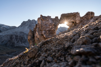 Eine natürliche Felsbrücke im Hochgebirge, durch deren steinerne Öffnung die tief stehende Sonne scheint, vor schroffen Berggipfeln