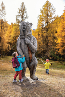 Wow, so gross ist ein Bär – das Modell des Bären steht am Wegrand des Senda da l’uors (Foto: © Nico Schaerer) Drei Kinder stehen um eine lebensgrosse Bärenstatue aus Arvenholz am Wegrand des Senda da l’uors