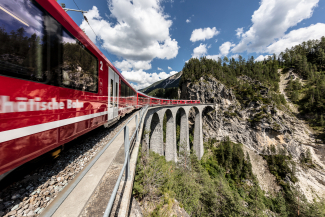 Ein roter Zug der Rhätischen Bahn fährt auf dem Landwasserviadukt in einer engen Rechtskurve über die hohen Steinpfeiler hinweg auf ein Tunnelportal in einer steilen Felswand zu