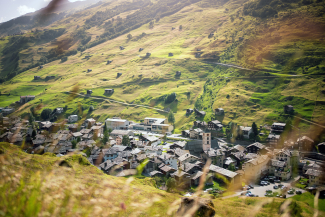Sunny mountain village of Vals: stone roofs and church tower in the valley, green slopes with scattered huts.