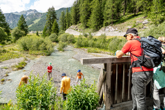 Eine Familie mit Kindern auf Kiesbänken am fliessenden Bergbach Rom im Val Müstair, im Vordergrund ein Wanderer auf einer Holzbrücke vor dichtem Nadelwald und Bergen