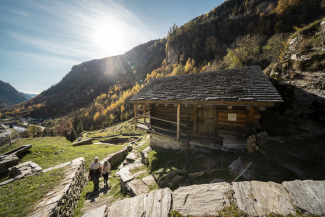Zwei Wanderer gehen über steinerne Stufen an einer traditionellen Holzhütte mit Steindach im sonnigen Val Calanca bergauf.