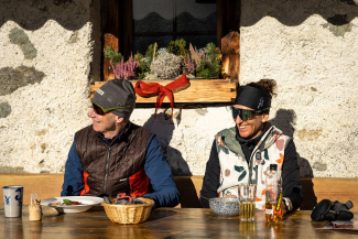 Zwei Langläufer sitzen lachend in der Sonne vor einer Berghütte beim Essen