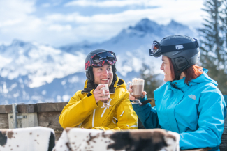 Roggis Baizli (Foto: © Tourismus Savognin Bivio Albula AG / Mattias Nutt) Zwei Skifahrerinnen geniessen heisse Getränke auf der Sonnenterrasse von Roggis Baizli mit Alpenpanorama im Hintergrund