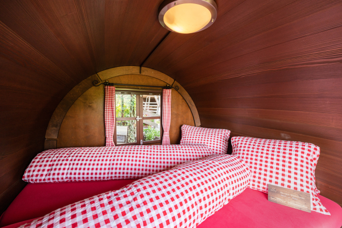 Gemütlicher Innenraum eines Schlaf-Fasses mit kariertem Bettbezug und Holzverkleidung