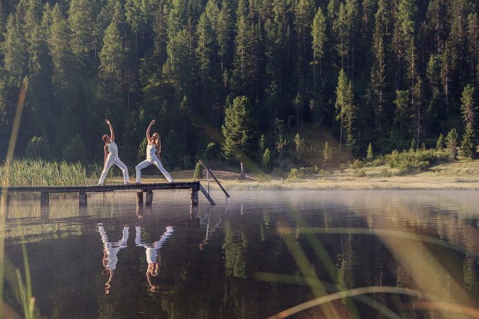 Mit natürlicher Entspannung den Alltag hinter sich lassen (Foto: © Urs Homberger) Zwei weiss gekleidete Frauen machen eine Yogaübung auf einem Holzsteg über einem Bergsee im Oberengadin