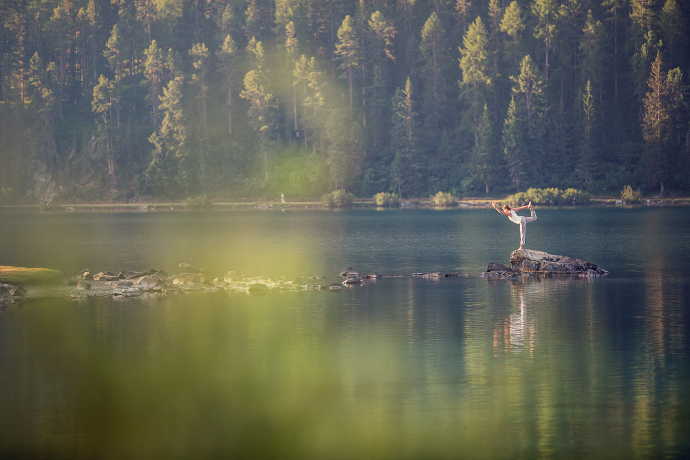 Abtauchen und die Sinne ins Gleichgewicht bringen im Oberengadin (Foto: © Engadin St. Moritz Tourismus / Filip Zuan) Eine Frau steht bei einer Yogaübung einbeinig auf einem grossen Stein in einem See im Oberengadin