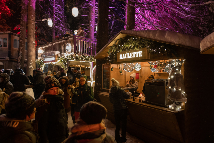 Besucherinnen und Besucher flanieren zwischen hell beleuchteten Marktständen im winterlichen Zauberwald Lenzerheide