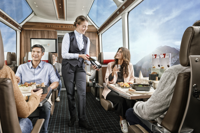 Glacier Express (Photo : © Glacier Express AG / Stefan Schlumpf) Une hôtesse de train montre à une femme une bouteille de vin dans le Glacier Express