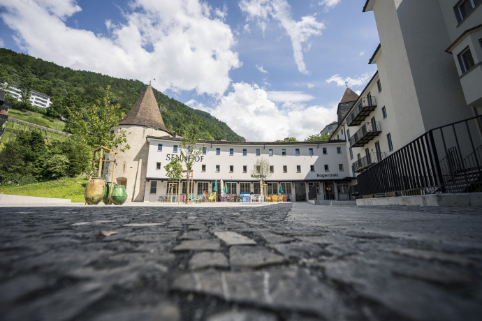 Aussenansicht des Bogentrakt Hostels im Sennhof Chur mit historischem Rundturm heller Fassade und bunter Terrasse vor grünem Berghintergrund