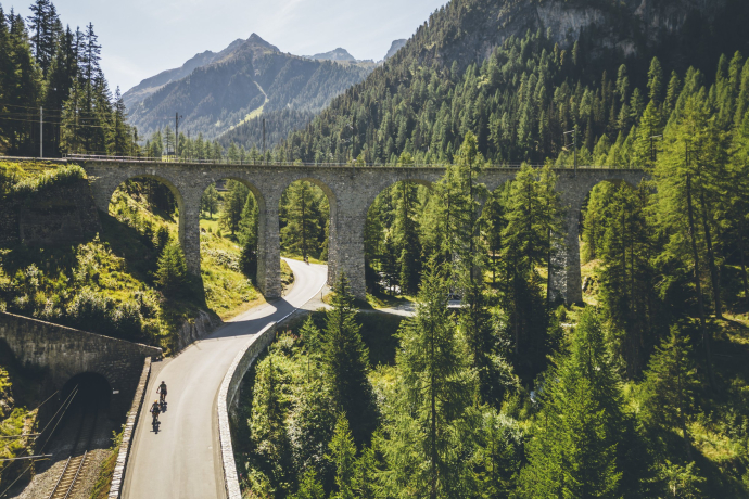 Zwei Velofahrer auf der Glacier Bike Tour unterfahren ein Bahnviadukt bei Bergün