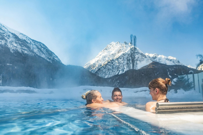 Three people relax in the outdoor pool of the Bogn Engiadina Scuol in the middle of a snowy winter landscape