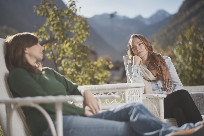 Achtsamkeit in der Natur – gemeinsames Innehalten mit Blick auf die Berge (Foto: © Graubünden Ferien / Samuel Trümpy) Zwei Frauen sitzen entspannt auf weissen Terrassenstühlen und unterhalten sich, im Hintergrund die Berge bei Bergün