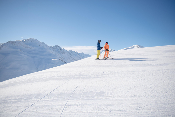 Zwei Skifahrer*innen stehen auf einer frisch präparierten Skipiste in der SkiArena Andermatt-Sedrun, umgeben von einer weiten Schneelandschaft und Berggipfeln unter blauem Himmel