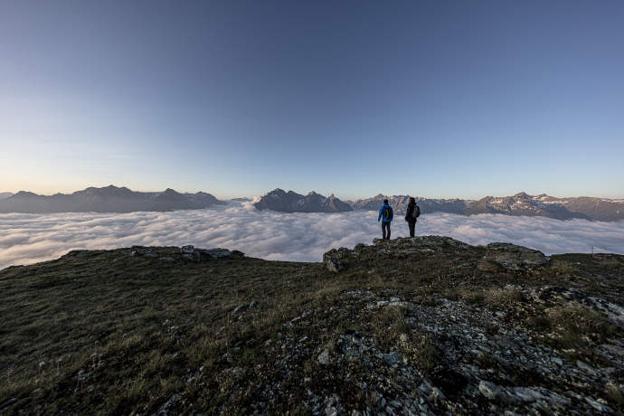 Two hikers standing on Mot da l’Hom above a sea of clouds with a view of the mountains in the Lower Engadine