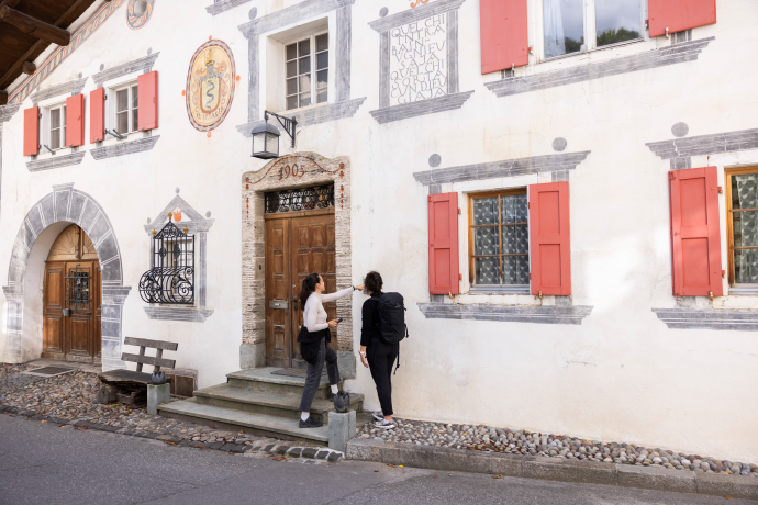Lors de la visite du village FiliTour à Filisur, les visiteurs découvrent le village à leur rythme, maison par maison (Photo : © Chemins de fer rhétiques / Nicola Pitaro) Deux femmes découvrent lors de la promenade FiliTour un bâtiment historique à Filisur avec des sgraffites