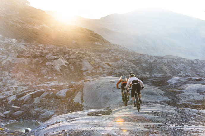 Zwei Mountainbikerinnen fahren bei Sonnenaufgang über felsiges Gelände in alpiner Landschaft
