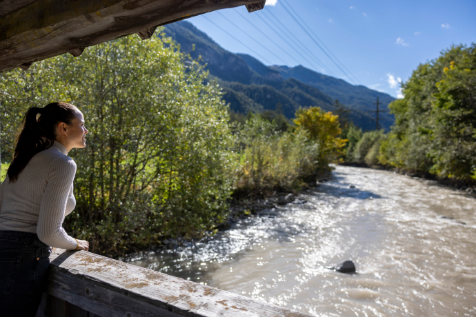 Le long du parcours aquatique Ansaina, de nombreux endroits invitent à s’arrêter, à observer et à s’émerveiller (Photo : © Chemins de fer rhétiques / Nicola Pitaro) Une femme regarde le fleuve depuis un pont en bois sur le chemin de l’Ansaina à Filisur