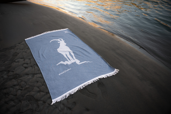 Stylisches «graubünden»-Badetuch aus SEAQUAL®-Garn – jetzt im Fanshop erhältlich (Foto: © Marco Hartmann) Badetuch in Eisblau mit weissem Steinbock-Motiv auf dunklem Sandstrand, nahe am Wasser gelegen