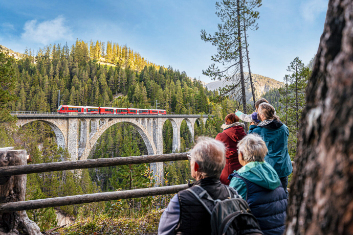 Gruppe von Menschen beobachtet einen roten Zug auf dem Landwasserviadukt von der Aussichtsplattform «Coray» aus inmitten eines Waldgebiets