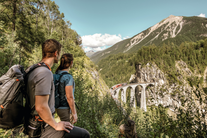 Aussichtsreicher Wanderweg bei Filisur mit Blick auf das Landwasserviadukt – ein beliebter Spot entlang der UNESCO-Strecke der Rhätischen Bahn (Foto: © Schweiz Tourismus / Rob Lewis) Zwei Wanderer blicken vom Aussichtspunkt bei Filisur auf das Landwasserviadukt und einen Zug der Rhätischen Bahn