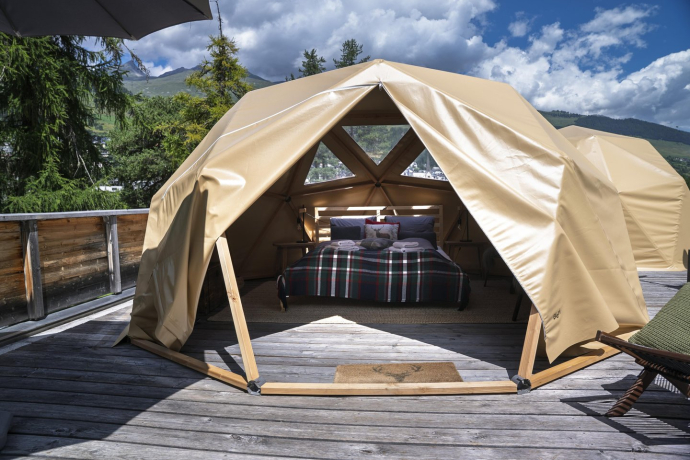 Geopods mit geoeffnetem Eingang und Blick auf ein Doppelbett mit karierter Decke, auf einer Holzterrasse in den Bergen.