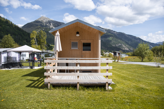 Kleine Holzhuette mit Terrasse und Sonnenschirm auf einer Wiese des Camping Calvaresc, umgeben von Bergen und blauem Himmel mit Wolken