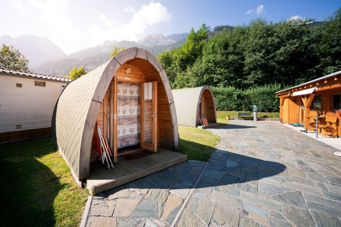 Mehrere halbrunde Holzhütten (Pods) auf dem Camping Calvaresc, mit Steinweg und Rasen, bei Sonnenschein und Bergen im Hintergrund