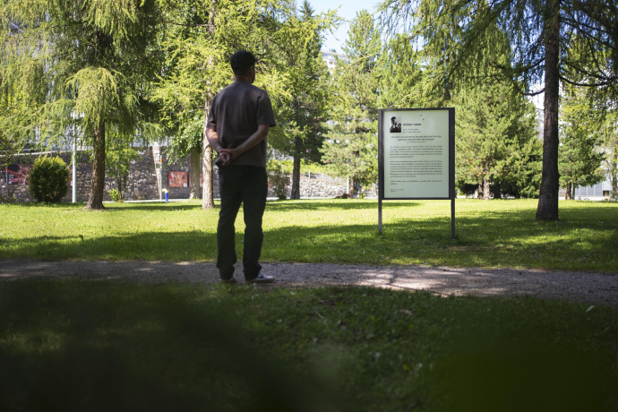 Eine Person betrachtet im Park von Davos Klosters eine Infotafel über Thomas Mann zwischen grünen Bäumen.