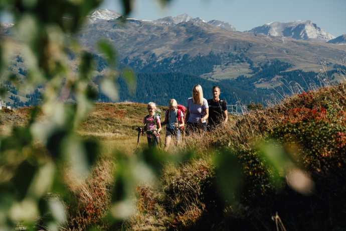 Eine Familie wandert auf einem Bergpfad, im Hintergrund hohe Berge und Wälder.