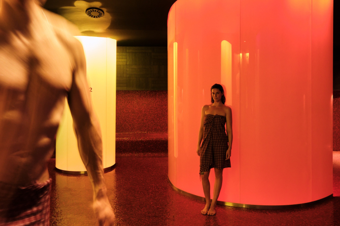 Stimmungsvolle Lichtspiele im Hamam des Hotel Castell in Zuoz – ein Ort der Ruhe und Wärme (Foto: © Yiorgos Kaplanidis, 2024) Frau im Hamam des Hotel Castell in Zuoz, angelehnt an eine rot beleuchtete Wand, im Vordergrund ein unscharfer Mann in Bewegung