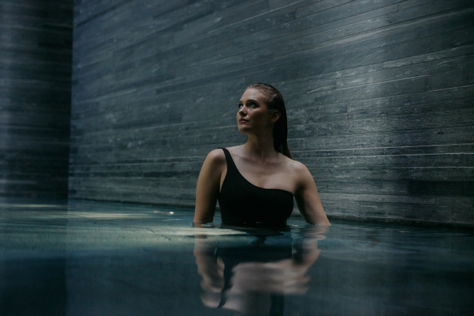 7132 Therme Vals – Wellness und Architektur vereint mit 30 Grad warmem, mineralisiertem Wasser aus der St. Petersquelle (Foto: © 7132 Hotel / Julien Balmer) Junge Frau im schwarzen Badeanzug steht im Innenbereich der 7132 Therme in Vals mit Steinwänden aus Valser Quarzit
