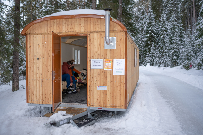 Into the skates and out into the winter adventure – the cosy changing huts at the start and end of the ice trail offer space to get ready and warm up (Photo: © Dominik Täuber / Tourismus Engadin Scuol Samnaun Val Müstair AG) Two people putting on their skates inside a wooden hut
