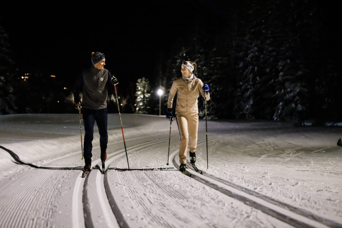Langläufer am nachlaufen (Foto: © Flilip Zuan) Zwei Langläufer gleiten bei Nacht auf einer beleuchteten Loipe durch einen verschneiten Wald. Der Himmel ist dunkel, und die Scheinwerfer werfen lange Schatten auf den Schnee.