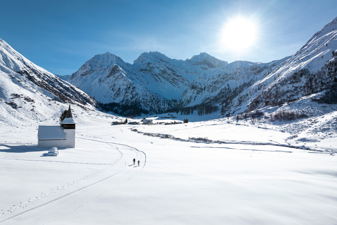 Langläufer auf der Loipe (Foto: © Flilip Zuan) Eine winterliche Landschaft in den Alpen, mit verschneiten Bergen im Hintergrund und einer kleinen Kirche im Vordergrund.