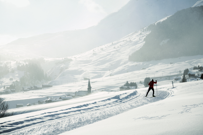 Langläufer auf der Loipe (Foto: © Schweiz Tourismus/Stefan Schlumpf) Eine Person fährt auf einer Langlaufloipe durch verschneite Landschaft. Im Hintergrund ist ein kleines Dorf mit einer Kirche und hohe Berge zu sehen, die von Nebel und Sonne durchzogen werden.