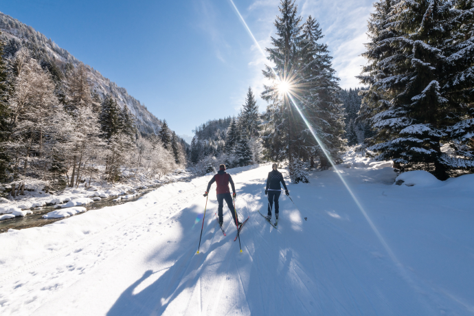 Langläufer auf der Loipe (Foto: © Mattias Nutt) Zwei Langläufer auf einer verschneiten Strecke, umgeben von winterlicher Berglandschaft und Bäumen, mit der Sonne im Hintergrund.