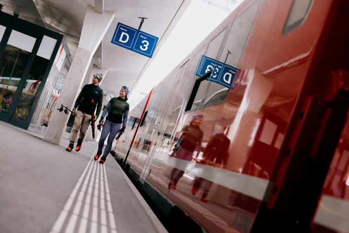 Perfekte ÖV Anbindungen von Loipe zu Loipe (Foto: © maloja Clothing GmBh) Ein Paar Langläufer geht auf einem Bahnhof im Engadin, mit der Rhätischen Bahn im Hintergrund.