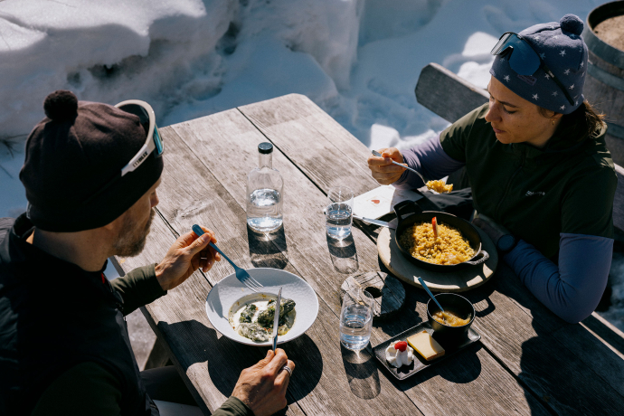 Sonnige Mittagspause im Restaurant (Foto: © Schweiz Tourismus / Nicola Fürer) Zwei Personen geniessen ein Mittagessen im Freien im Schnee, auf einem Holztisch, mit einem Teller und Getränken vor ihnen.