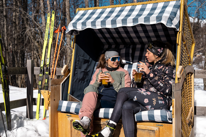 Zwei Langläuferinnen geniessen eine Pause in einem Strandkorb (Foto: © Filip Zuan) Zwei Frauen sitzen in einem Strandkorb in den Bergen, während sie eine Pause vom Langlaufen machen. Sie tragen sportliche Winterbekleidung und geniessen ihre Getränke, umgeben von verschneiter Landschaft.