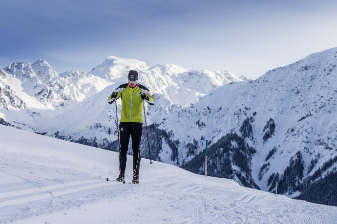 Langläufer auf einer verschneiten Loipe mit beeindruckendem Bergpanorama im Hintergrund, in der Region Unterengadin, während er die Spur auf der präparierten Strecke entlangfährt.