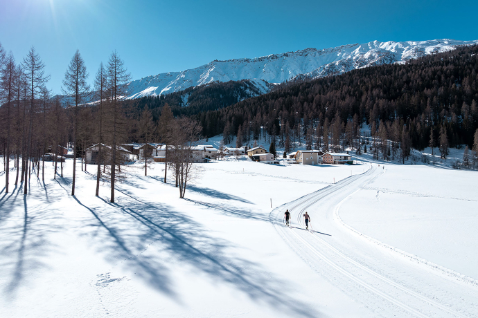Zwei Langläufer auf einer Loipe im Unterengadin (Foto: © Filip Zuan) Das Bild zeigt eine winterliche Landschaft im Unterengadin mit Langläufern auf einer Loipe. Im Hintergrund sieht man verschneite Hügel und eine kleine, traditionelle Bergsiedlung. Klare, sonnige Wetterbedingungen.