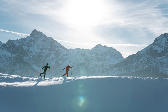 Klassische Langläufer vor wunderschönem Bergpanorama (Foto: © Filip Zuan) Zwei Langläufer unterwegs auf einer Loipe im Engadin, mit den majestätischen Bergen im Hintergrund und einer winterlichen Landschaft unter strahlendem Sonnenschein.