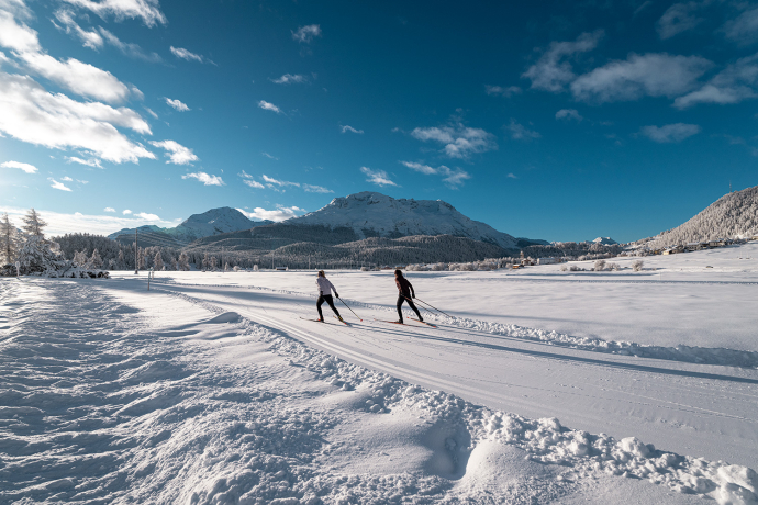 Das Oberengadin begeistert mit seiner endlosen Weite, der glitzernden Seenplatte und perfekt präparierten Loipen – ein Paradies für Langläuferinnen und Langläufer (Foto: © Filip Zuan) Zwei Langläufer auf weiter, sonniger Loipe mit verschneiten Bergen und blauen Himmel im Oberengadin