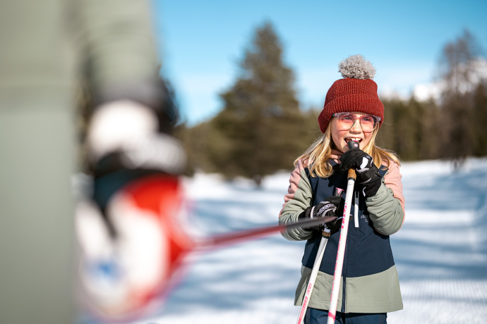 So macht Wintersport Spass: Die Langlaufregion Graubünden begeistert Gross und Klein mit Sonne, Schnee und Freude in Bewegung (Foto: © Filip Zuan) Lachendes Mädchen mit Mütze und Langlaufstöcken auf sonniger Loipe
